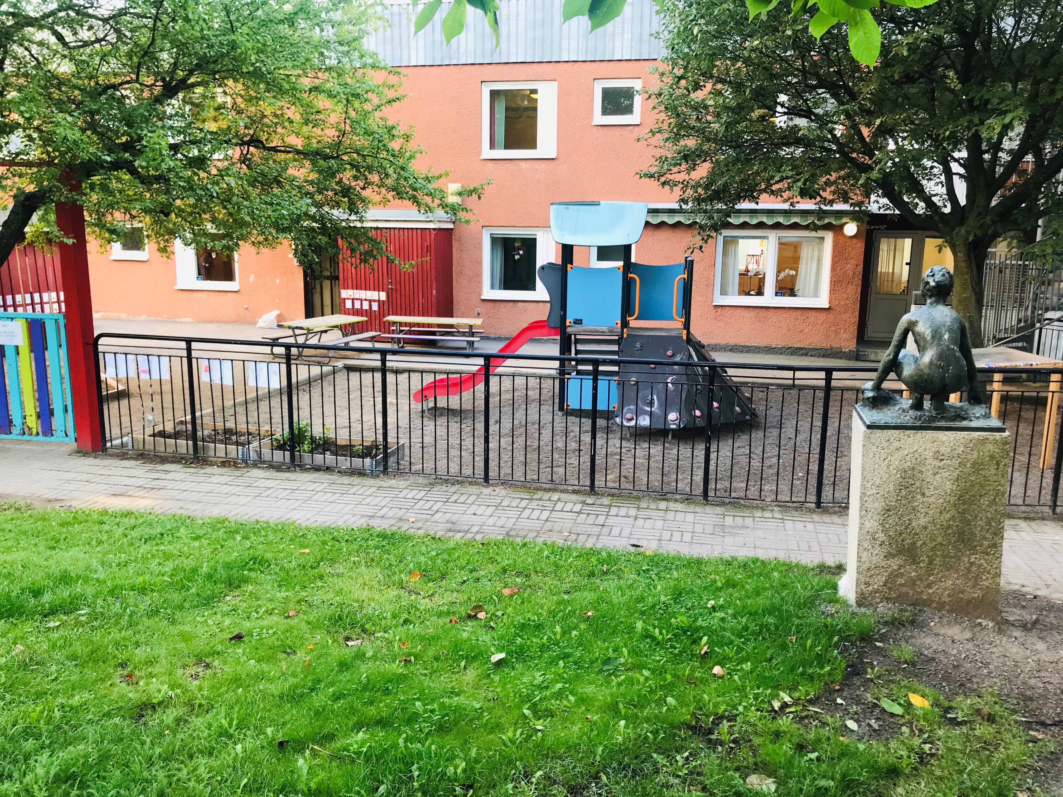 En röd tegelbyggnad med gård. På gården finns en klätterställning. Framför gården finns ett svart staket med röd grind.