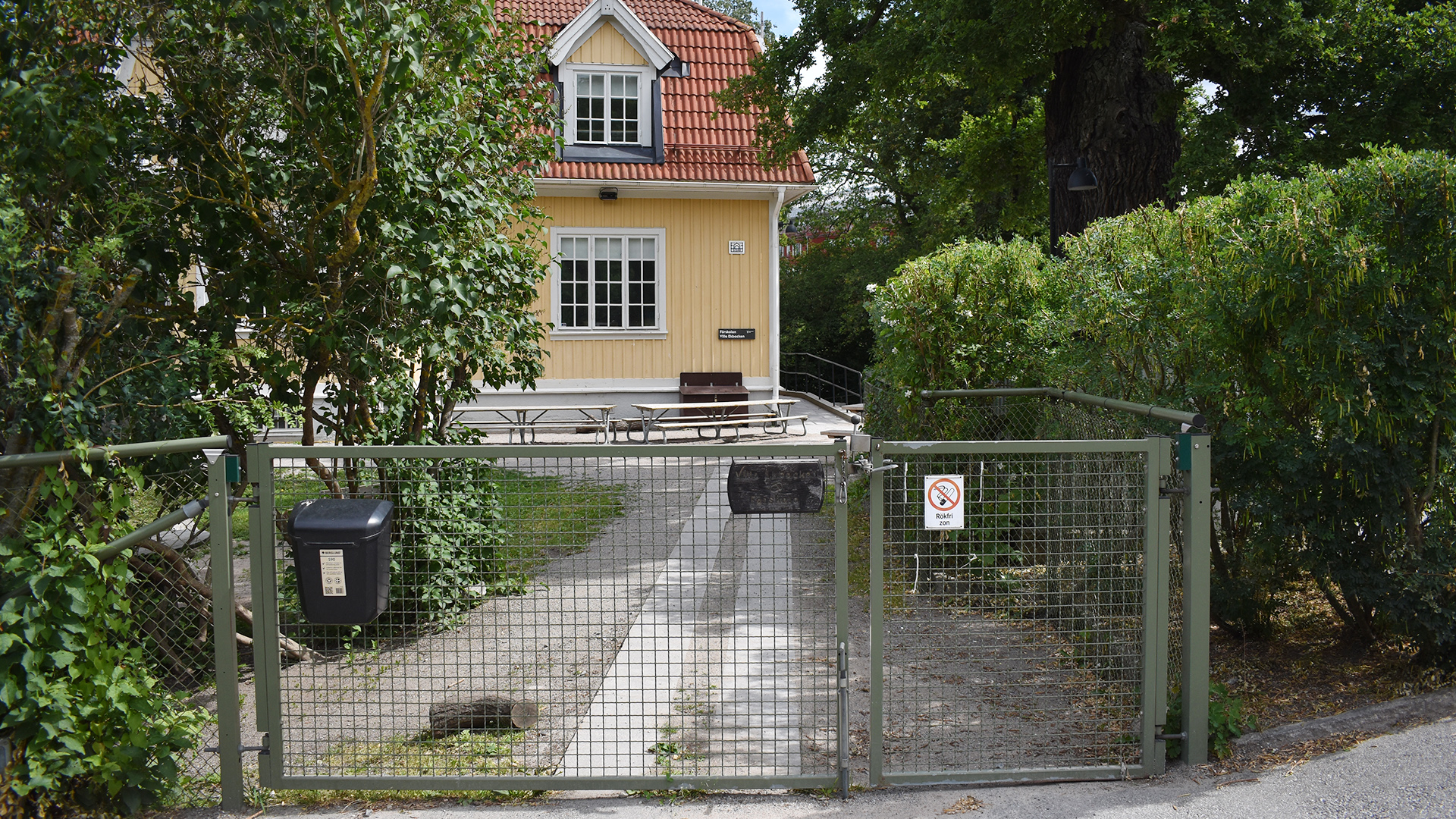 En gul villa med vita knutar, omgiven av träd och höga buskar. I förgrunden en grind.