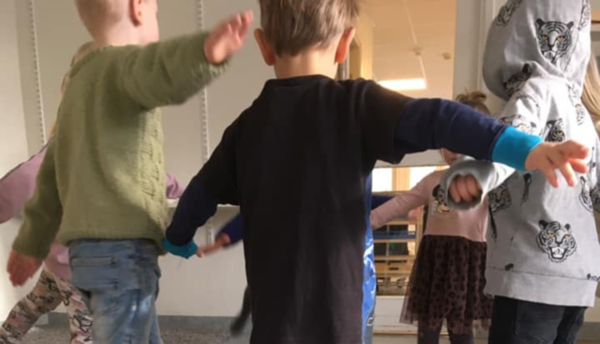En grupp förskolebarn leker i en cirkel inomhus och håller varandras händer.