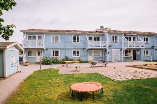 Ljusblått trähus i två våningar och en skolgård med sandlåda och sittplatser.