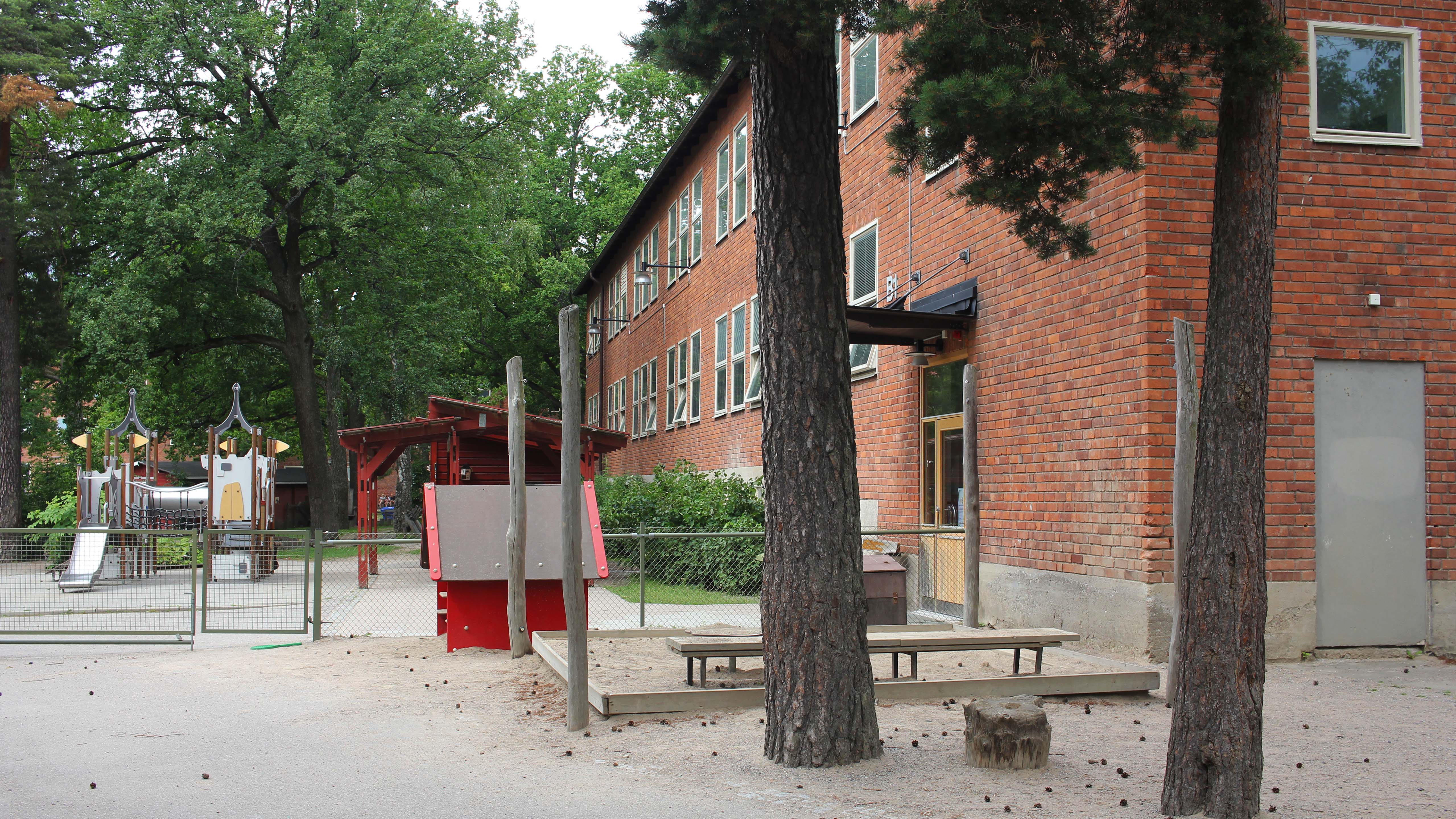Ett flerfamiljshus i rött tegel med en inhägnad gård med lövträd och buskar. På gården syns en klätterställning i metall, gult och rött. Utanför den gröna inhägnaden finns två tallar, ett rött lekhus och en sandlåda.