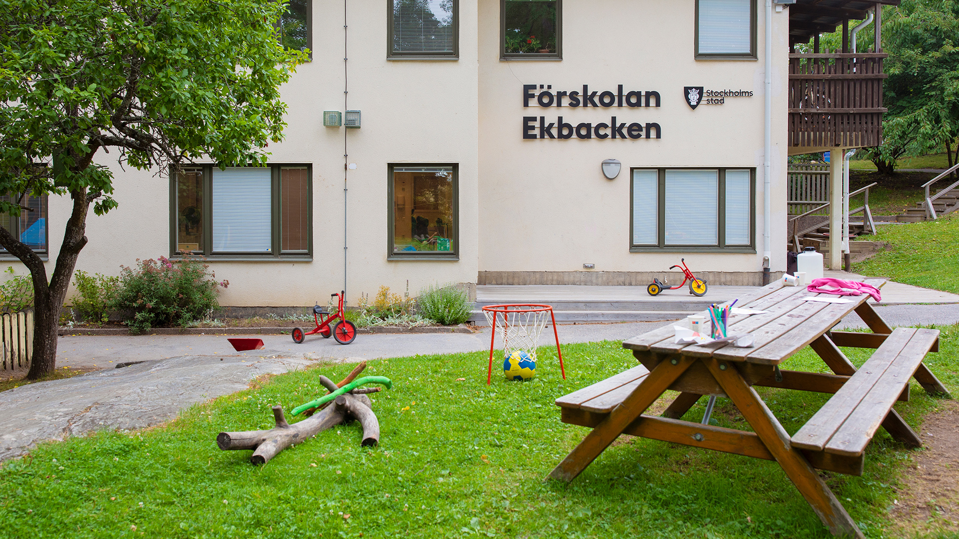 Förskolans gård med träd, planteringar, asfalt med trehjulingar och gräsmatta med leksaker och pysselbord.