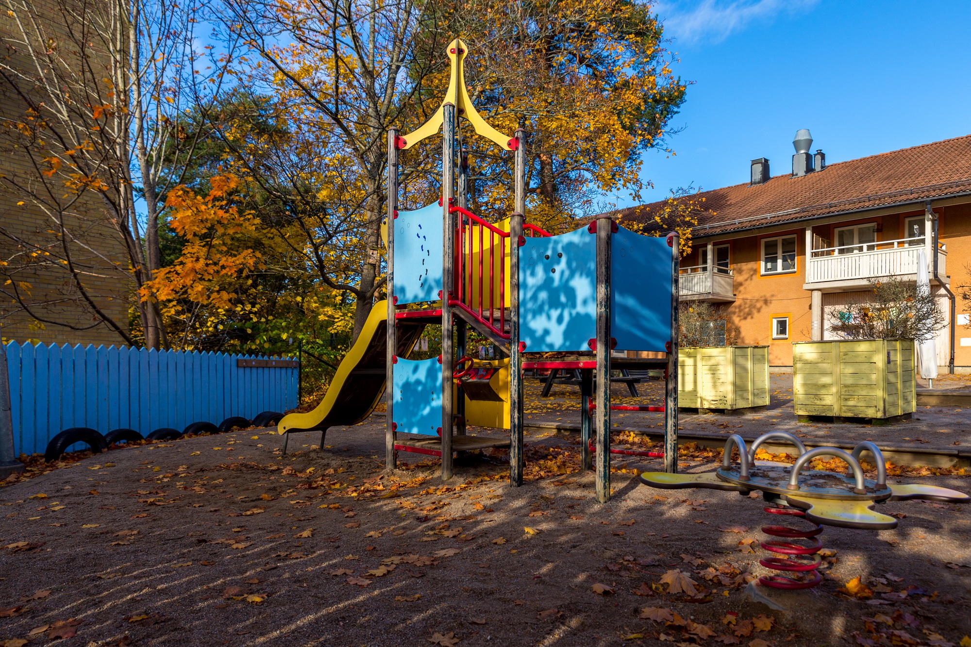 Gård med klätterställning, sandlåda och träd framför en byggnad