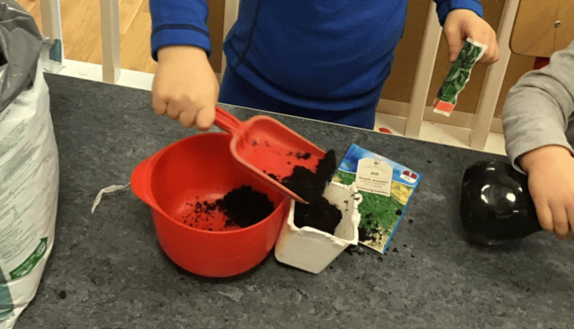 Förskolebarn planterar frön inomhus med jord, skopa och krukor på ett bord.