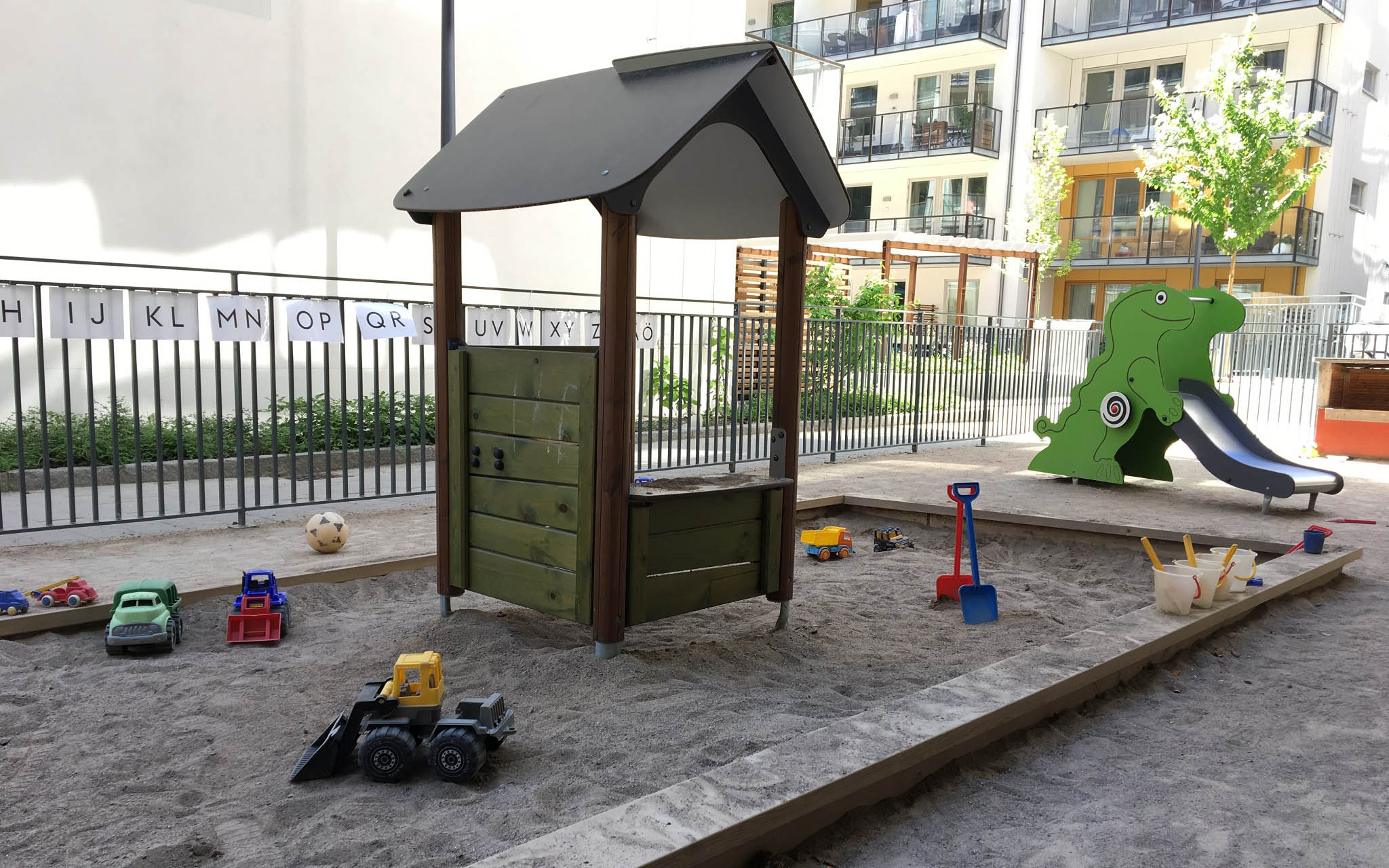 Utemiljö med lekytor, sandlåda och rutschkana. Ljusa flerbostadshus i bakgrunden.