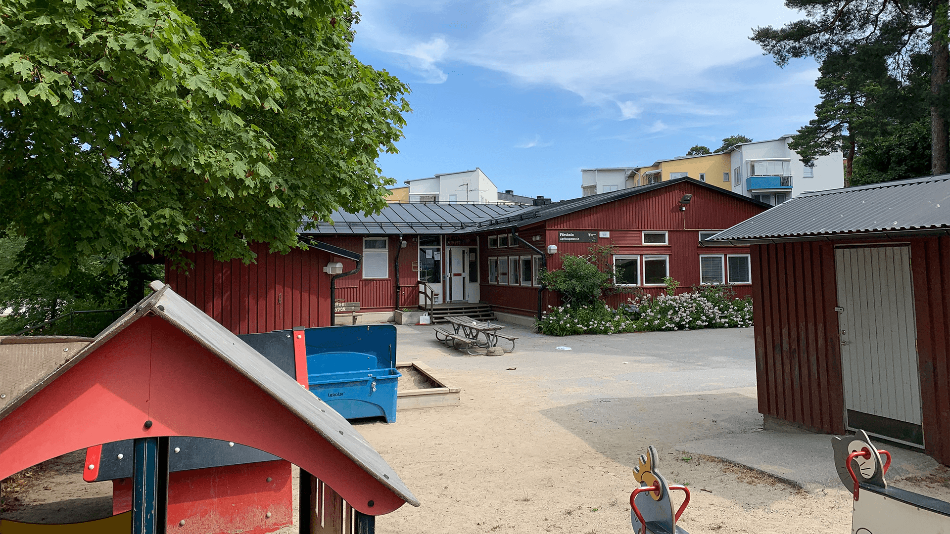 Stor gård med träd och bänkar. I förgrunden skymtar lekredskap. I bakgrunden förskolebyggnaden.