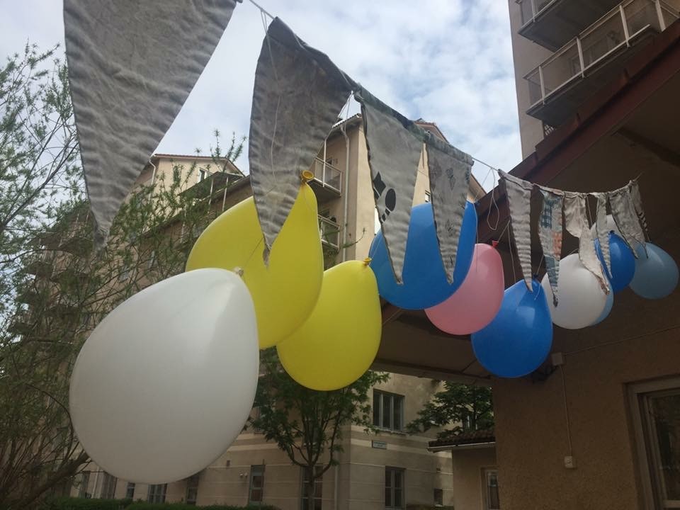 Blå, gula, rosa och vita ballonger och vita vimplar med tryck hänger utanför flerfamiljshus.