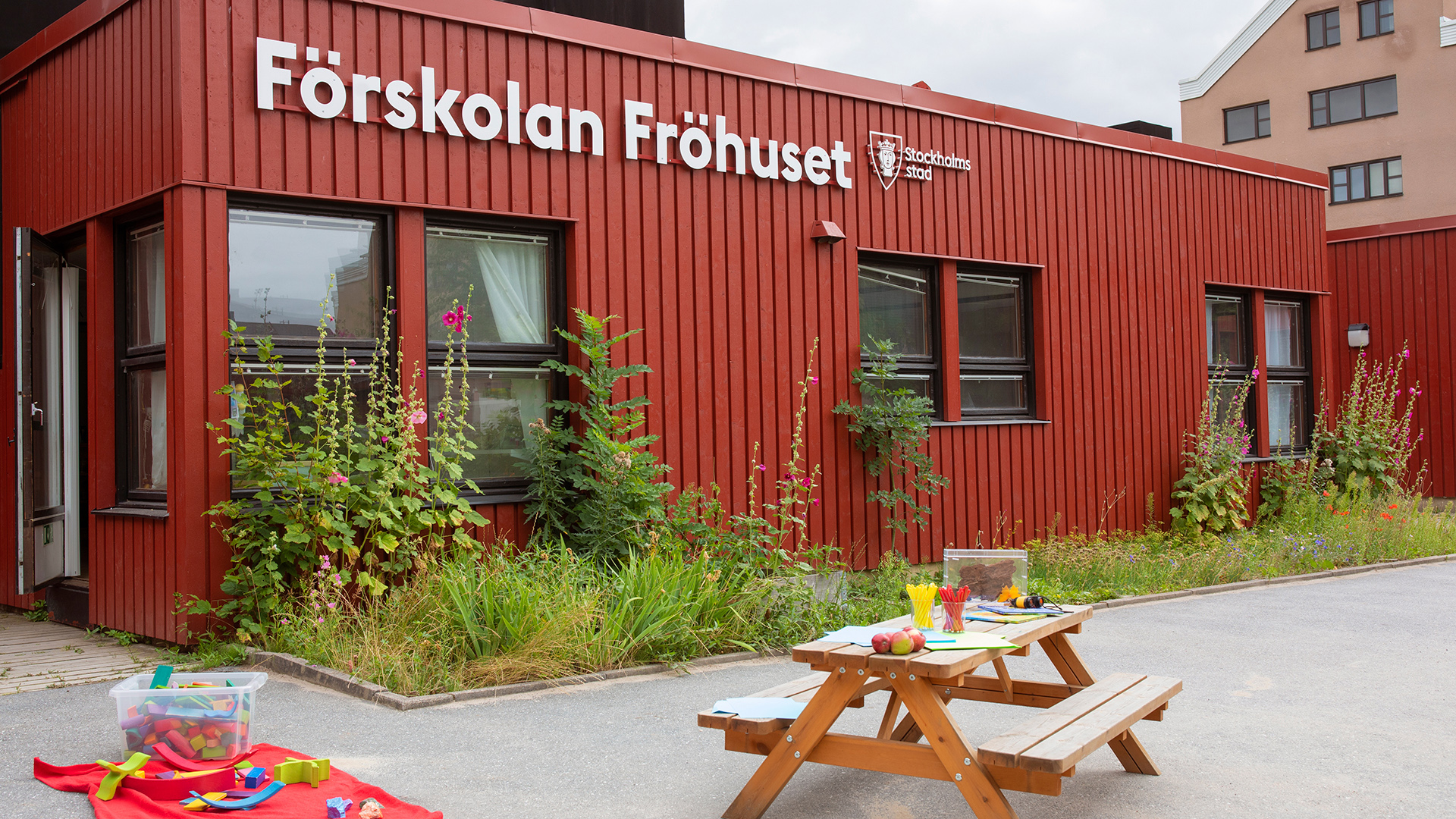 Förskolegård med planterade buskar och blommor i rabatten. I övrigt asfalt med framtagna leksaker och bord med pyssel.