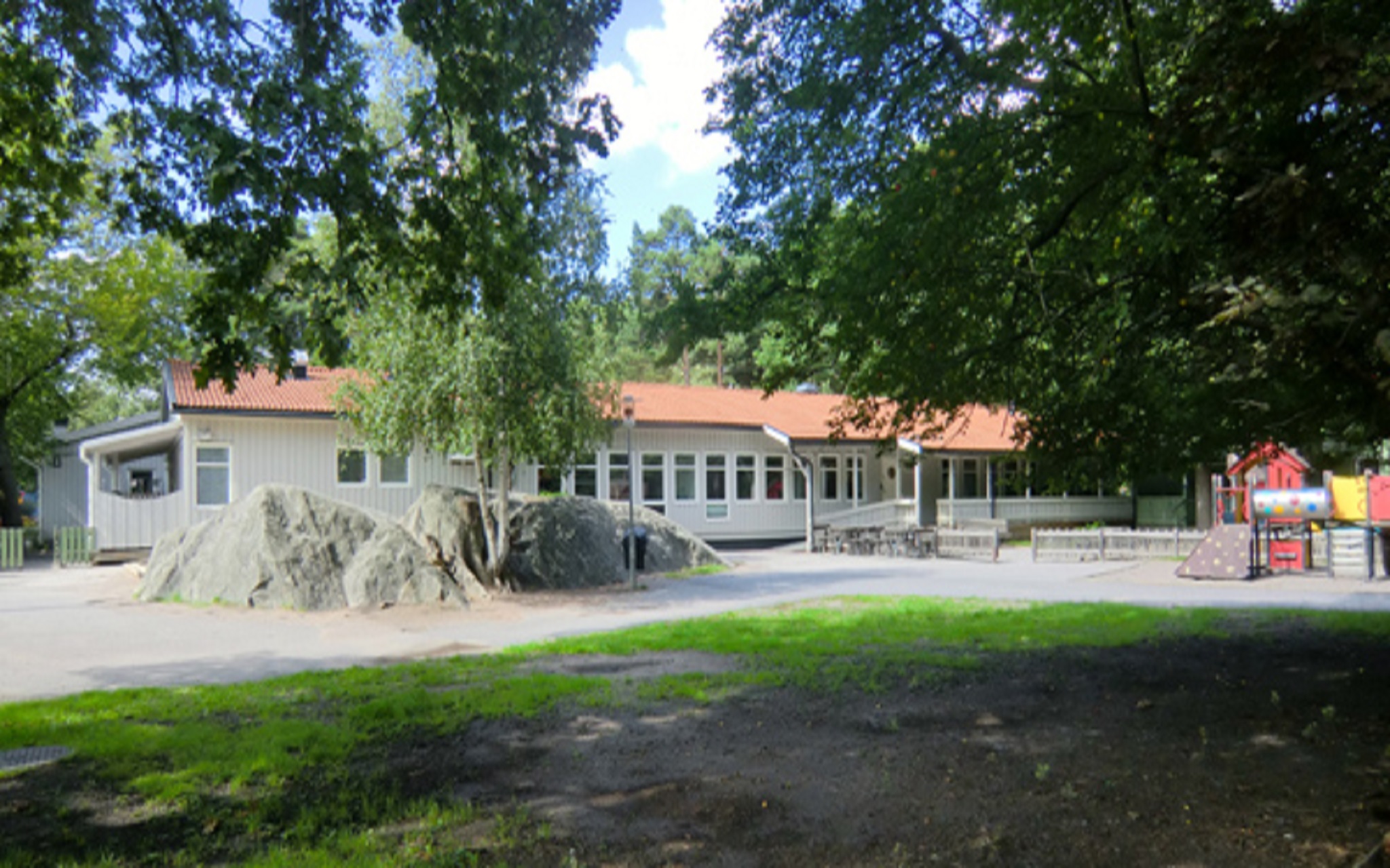 Vit enplansbyggnad med gård med grönska, litet berg och klätterställning.