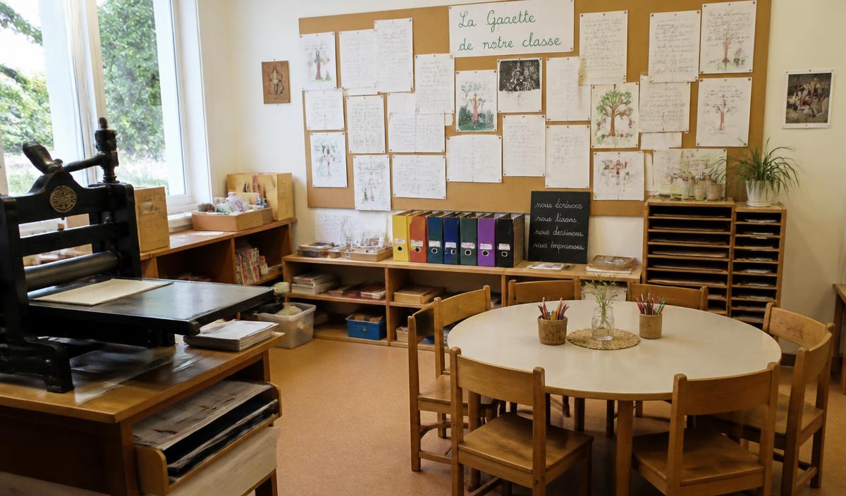 En freinetinspirerad förskolehörna med ett litet tryckpress, barnens handskrivna tidning på anslagstavlan och ett runt bord för samarbete